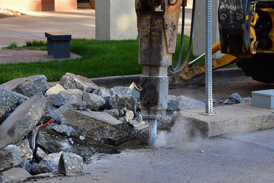 A tractor with a concrete breaker demolishing a sidewalk. 